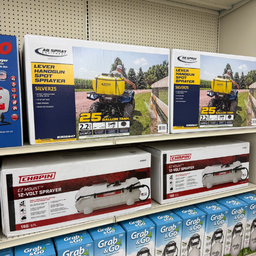 Shelves displaying various agricultural sprayers in boxes. Top shelf has yellow lever handgun spot sprayers, bottom shelf features 12-volt sprayers.