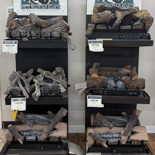 Display of six fireplace log sets on shelves, labeled with prices ranging from $630 to $1142. The logs appear realistic and varied in texture.