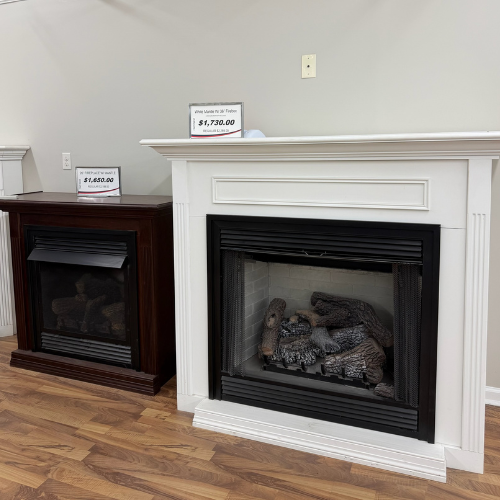 Two electric fireplaces are displayed side by side on a wooden floor. The left is dark wood priced at $1,620.00, and the right is white priced at $1,730.00.