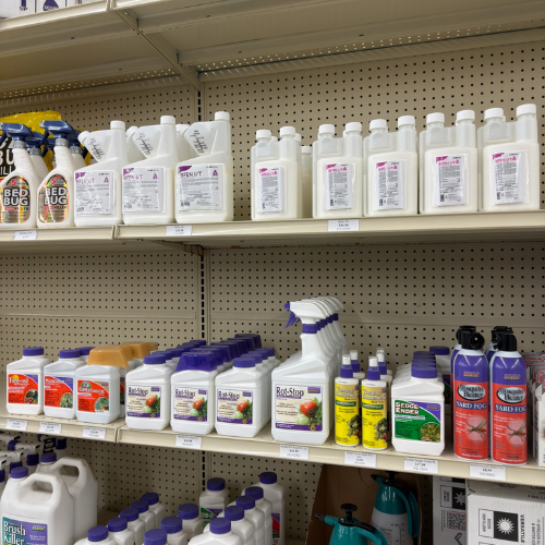 Shelves stocked with various garden and pest control products, including sprays and containers. Labels feature colorful graphics and text. Brightly lit store aisle.