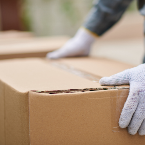 Gloved hands carefully handle a large cardboard box sealed with tape, conveying a sense of care and precision in moving or shipping items.